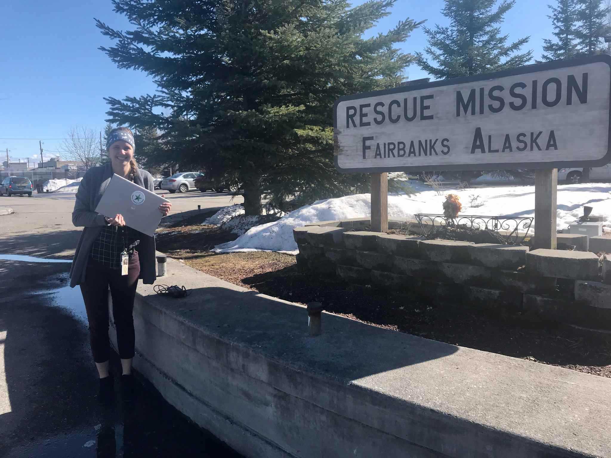 A woman holds up a laptop with a Green Star sticker in front of the Rescue Mission of Fairbanks Alaska outside on a sunny day